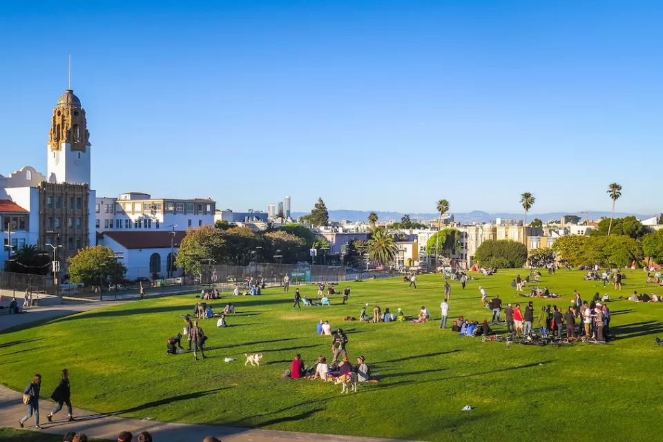 View of people sitting in lightly sloped green lawn with Spanish colonial-style building and cityscape in background in San Francisco, California.
